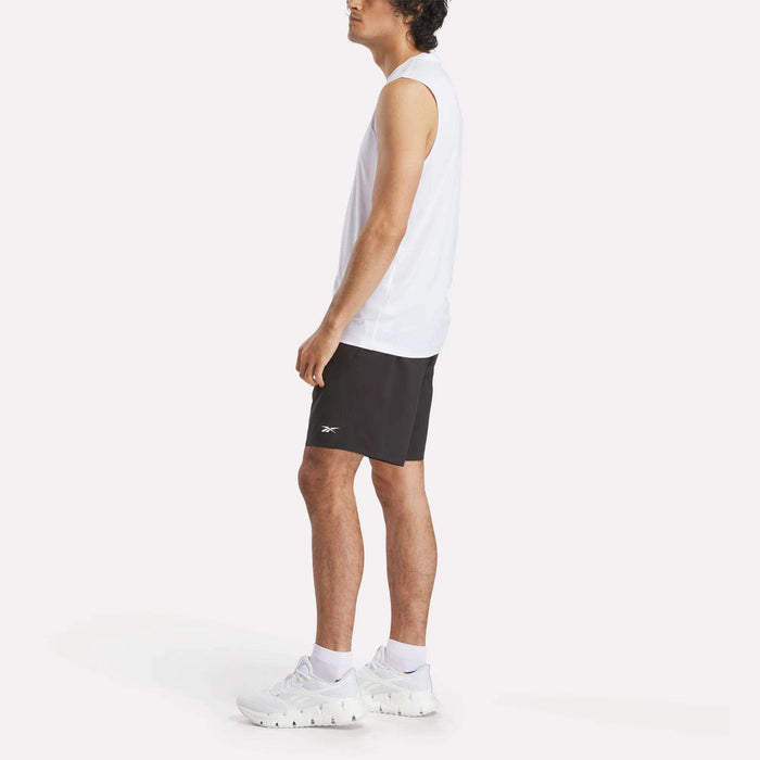A man stands sideways against a plain white background, wearing a Men's Sleeveless Tech Training T-Shirt by Reebok, black shorts, and white athletic shoes.