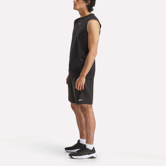 A man wears the Reebok Men's Train Sleeveless Tech Tee, black shorts, white socks, and black sneakers while standing in profile against a plain white background.
