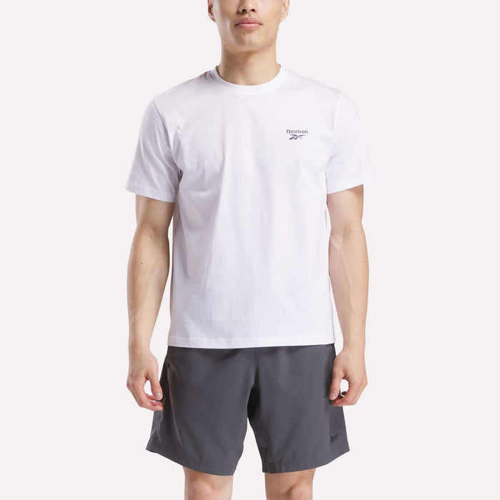 A man faces forward wearing a Reebok Men's Small Logo Crewneck Short Sleeve T-Shirt in white, made from soft cotton jersey, paired with dark gray shorts against a plain light background.