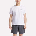 A man faces forward wearing a Reebok Men's Small Logo Crewneck Short Sleeve T-Shirt in white, made from soft cotton jersey, paired with dark gray shorts against a plain light background.