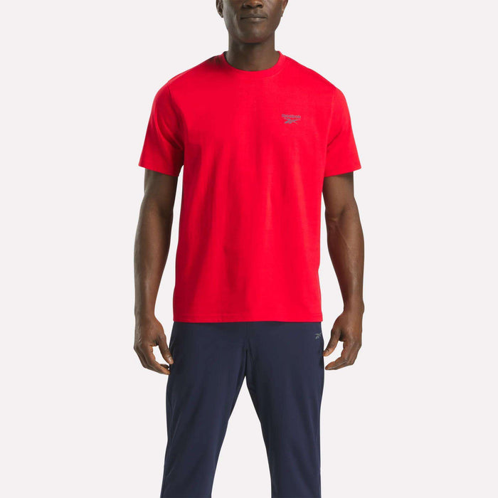 A person stands facing forward, wearing a bright red Reebok Men's Small Logo Crewneck Short Sleeve T-Shirt and dark blue pants against a plain white background.