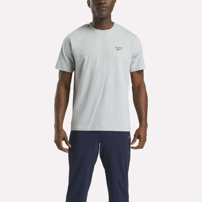 A man faces forward, wearing a Reebok Men's Small Logo Crewneck Short Sleeve T-Shirt in light grey cotton jersey and dark navy pants, set against a plain white background.