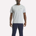 A man faces forward, wearing a Reebok Men's Small Logo Crewneck Short Sleeve T-Shirt in light grey cotton jersey and dark navy pants, set against a plain white background.