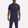 A man in a navy blue Reebok Men's Small Logo Crewneck Short Sleeve T-Shirt stands facing the camera against a plain white background. This classic fit tee features a small Reebok logo on the left chest.