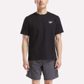 A man in a Reebok Men's Small Logo Crewneck Short Sleeve T-Shirt, black cotton jersey with a small white logo on the chest, paired with gray shorts, stands against a plain white background.