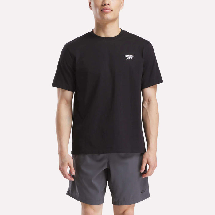 A man in a Reebok Men's Small Logo Crewneck Short Sleeve T-Shirt, black cotton jersey with a small white logo on the chest, paired with gray shorts, stands against a plain white background.