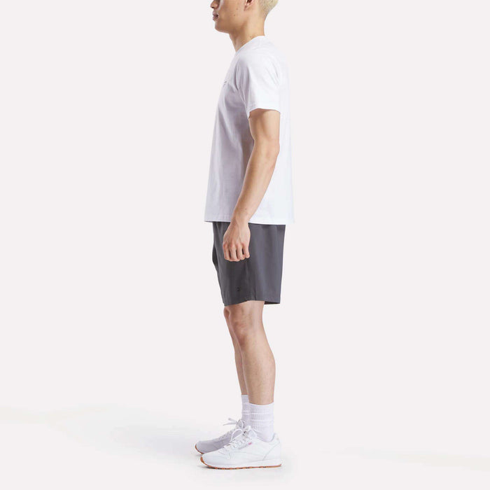 A person stands in profile facing left, wearing a Reebok Men's Small Logo Crewneck Short Sleeve T-Shirt, dark gray shorts, white socks, and white Reebok athletic shoes against a plain light background.