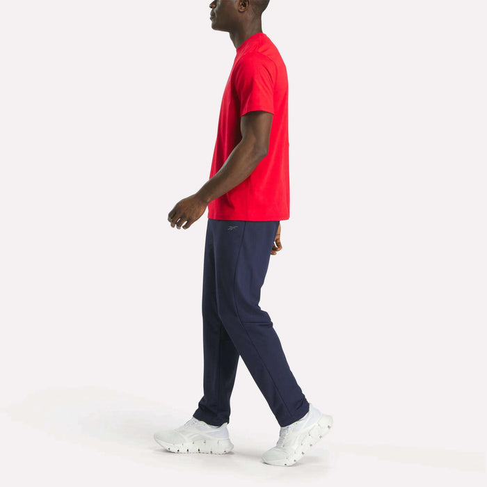 A man walks left, wearing a bright red Reebok Men's Small Logo Crewneck Short Sleeve T-Shirt in soft cotton jersey, dark blue pants, and white sneakers. The background is plain white.
