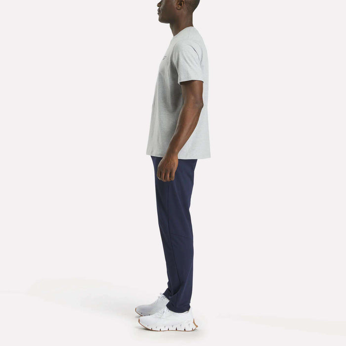 A man stands in profile facing left, wearing a Reebok Men's Small Logo Crewneck Short Sleeve T-Shirt in light gray cotton jersey, paired with navy blue athletic pants and white sneakers, against a plain light background.