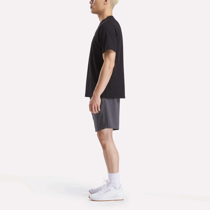 A man stands in profile against a white background, wearing a Reebok Men's Small Logo Crewneck Short Sleeve T-Shirt in black, gray shorts, white crew socks, and white athletic shoes.