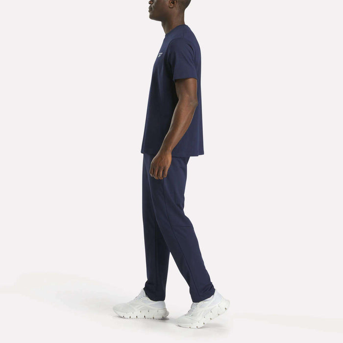 A man stands in profile against a white background, wearing a Reebok Men's Small Logo Crewneck Short Sleeve T-Shirt in navy blue cotton jersey, paired with matching navy pants and white athletic sneakers.