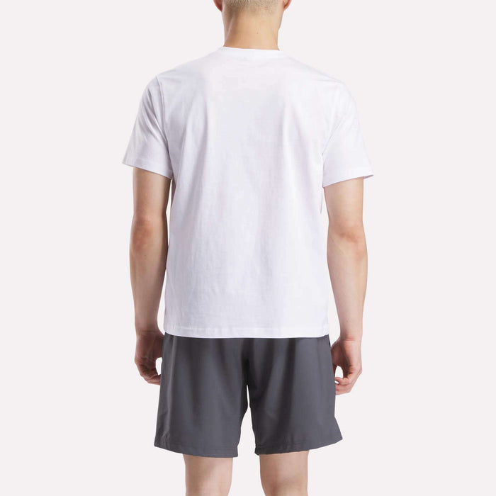 A person stands with their back to the camera, wearing a Reebok Men's Small Logo Crewneck Short Sleeve T-Shirt in white and dark gray shorts against a light background.