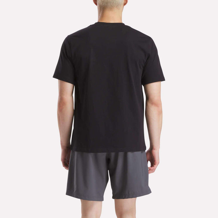 A person with short blonde hair faces away, wearing a plain black Reebok Men's Small Logo Crewneck Short Sleeve T-Shirt and dark gray shorts against a light background.