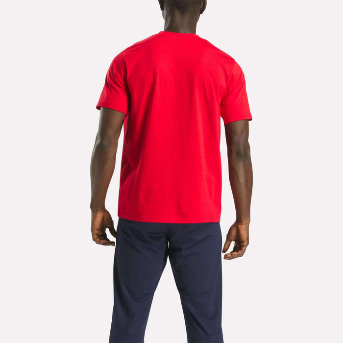 A person stands facing away in a bright red Reebok Men's Small Logo Crewneck Short Sleeve T-Shirt made of soft cotton jersey and dark blue pants, set against a plain light background.
