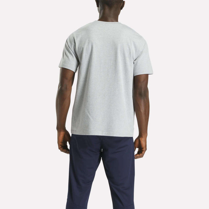 A person stands facing away, wearing the Reebok Men's Small Logo Crewneck Short Sleeve T-Shirt in light gray and dark navy pants, set against a plain white background.