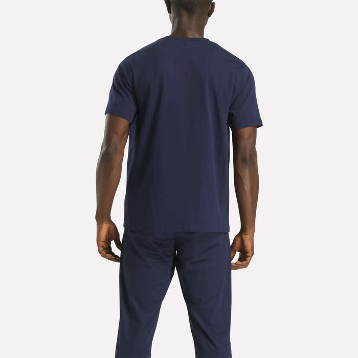 A person stands with their back to the camera, wearing a navy blue Reebok Men's Small Logo Crewneck Short Sleeve T-Shirt and matching pants against a plain light background.