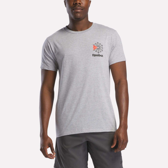 A man stands against a plain white background wearing a light gray Reebok Men's Starcrest Logo Graphic T-Shirt with a small logo on the left chest, paired with dark gray shorts.