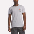 A man stands against a plain white background wearing a light gray Reebok Men's Starcrest Logo Graphic T-Shirt with a small logo on the left chest, paired with dark gray shorts.
