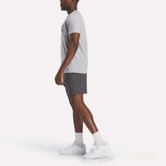 A man stands in profile against a white background wearing a light gray Reebok Men's Starcrest Logo Graphic T-Shirt, dark gray athletic shorts, white crew socks, and white Reebok sneakers.