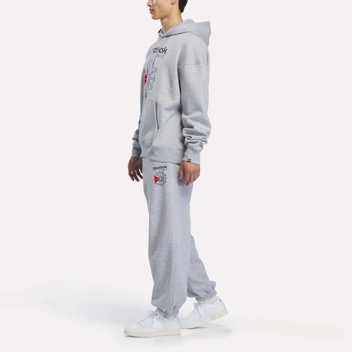 A person wearing a gray Reebok Men’s Starcrest Relaxed Hoodie made from brushed-back fleece, paired with matching sweatpants and white sneakers, stands in profile against a plain white background.
