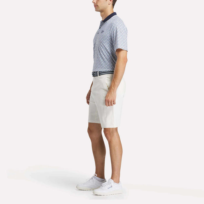 A man in profile wears a Reebok Men's Sublimation Print Shirt, white knee-length shorts, a striped belt, and white sneakers against a plain light background.
