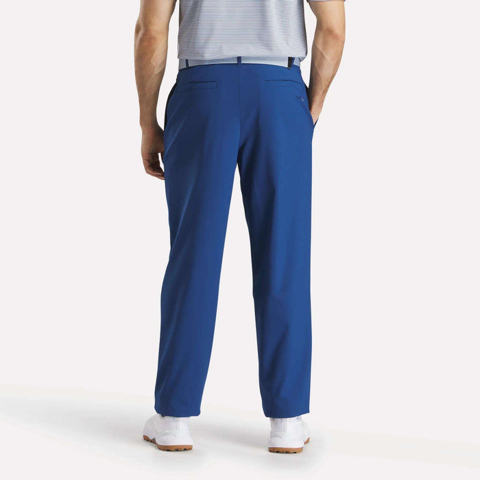 A man faces away in Reebok Men's The Favorite Golf Pants - Relaxed Fit (bright blue), paired with a tucked-in light gray striped shirt and white sneakers. His hands are in his pockets against a plain white background.