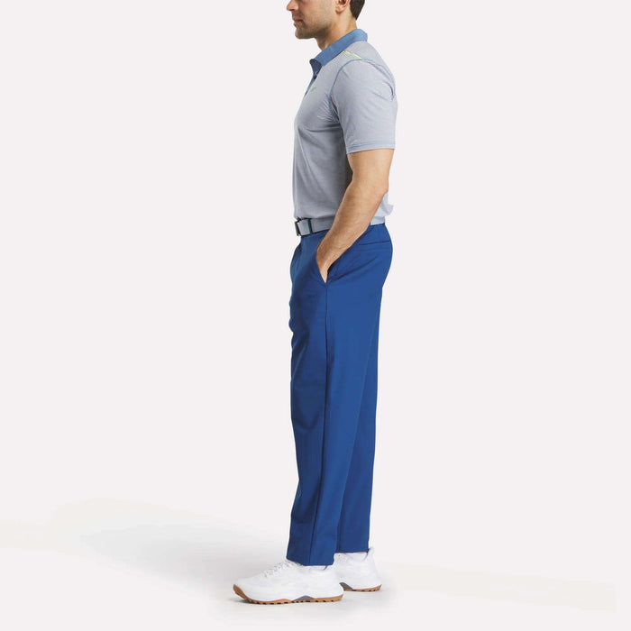 A man stands in profile wearing Reebok Men's The Favorite Golf Pants - Relaxed Fit in bright blue, paired with a light gray polo, black belt, and white sneakers against a plain white background.