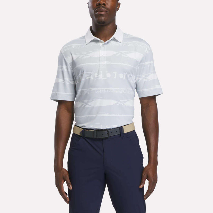A man wears the Reebok Men's Tournament Polo Shirt in Grey/White, styled with navy blue pants and a beige belt, standing against a plain white background.