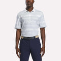 A man wears the Reebok Men's Tournament Polo Shirt in Grey/White, styled with navy blue pants and a beige belt, standing against a plain white background.