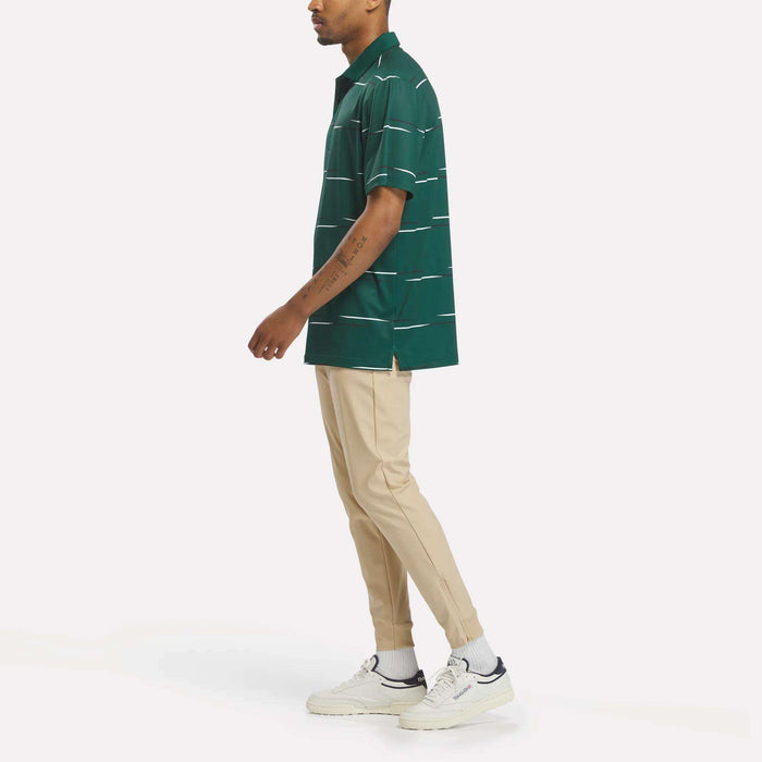 A person stands sideways on a white background wearing the Reebok Men's Tournament Polo Shirt in green stripes, paired with beige pants, white socks, and white sneakers.