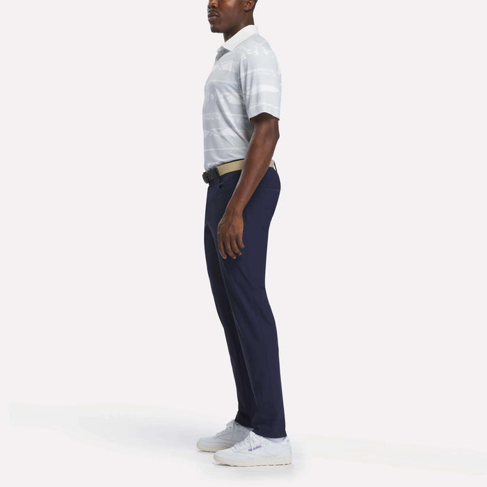 A man stands in profile against a plain white background, showcasing a tournament-ready look in the Reebok Men's Tournament Polo Shirt with light gray stripes, paired with navy blue pants, white sneakers, and a beige belt.