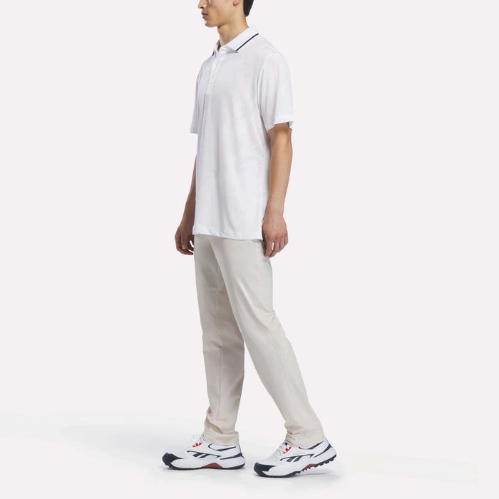 A man stands against a plain white background wearing a white Reebok Men’s Tournament Polo Shirt, light beige pants, and white athletic shoes with dark accents.