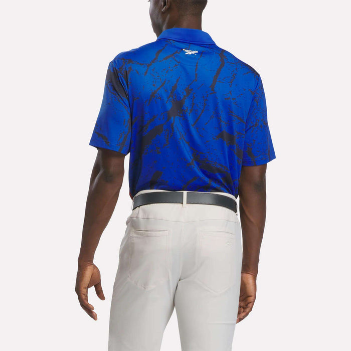 A man is seen from the back, standing against a plain white background, wearing the Reebok Men's Tournament Polo Shirt in a blue and black pattern and light gray pants.