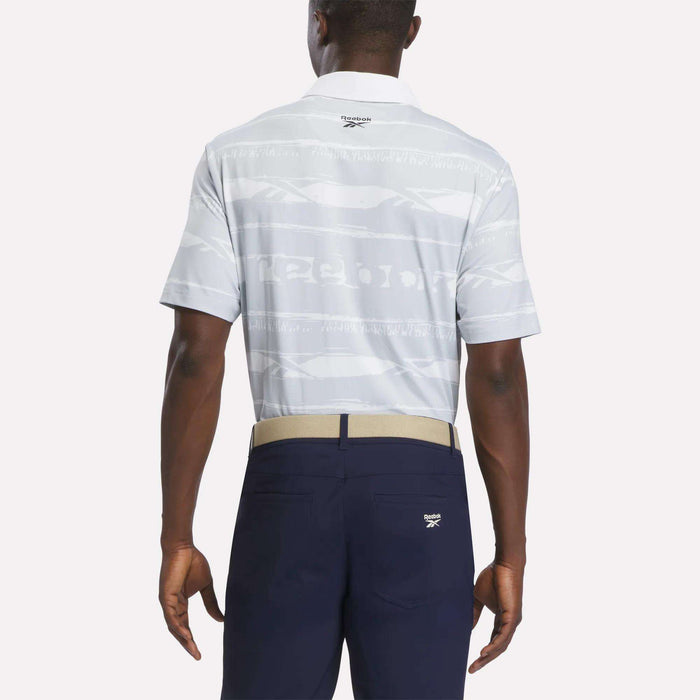 A man is seen from the back wearing the Reebok Men's Tournament Polo Shirt in light gray with a subtle pattern, paired with navy blue pants and a beige belt. Set against a plain light background, his outfit seamlessly blends classic style and modern performance—perfect for tournament-ready looks.