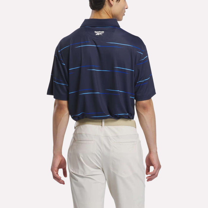 A man stands facing away against a plain white background, wearing the Reebok Men’s Tournament Polo Shirt in navy blue with thin blue stripes and light-colored pants—a subtle nod to classic golf polo style.