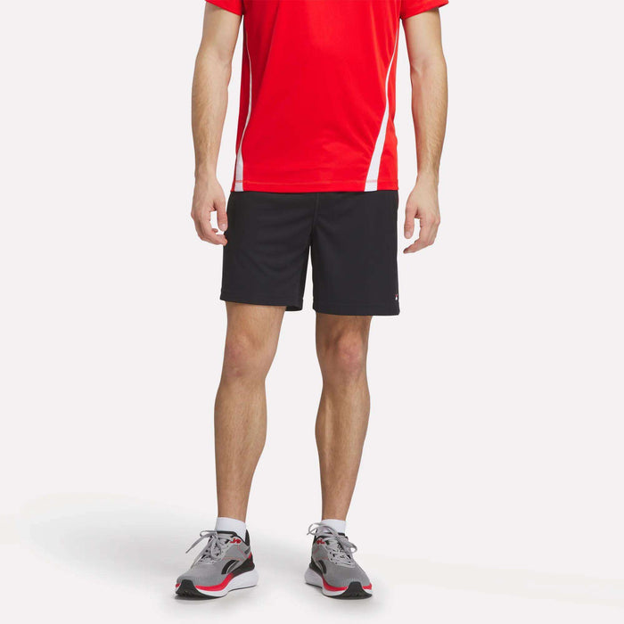 A person wearing a red short-sleeve athletic shirt, Reebok Men's Train Blocked Knit Shorts in black, and gray running shoes stands against a plain white background. The image is cropped just below the shoulders.