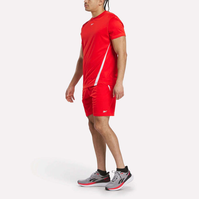 A person stands on a plain light background wearing bright red Reebok Men's Train Blocked Knit Shorts with a matching T-shirt, both accented in white, and gray, black, and red running shoes.