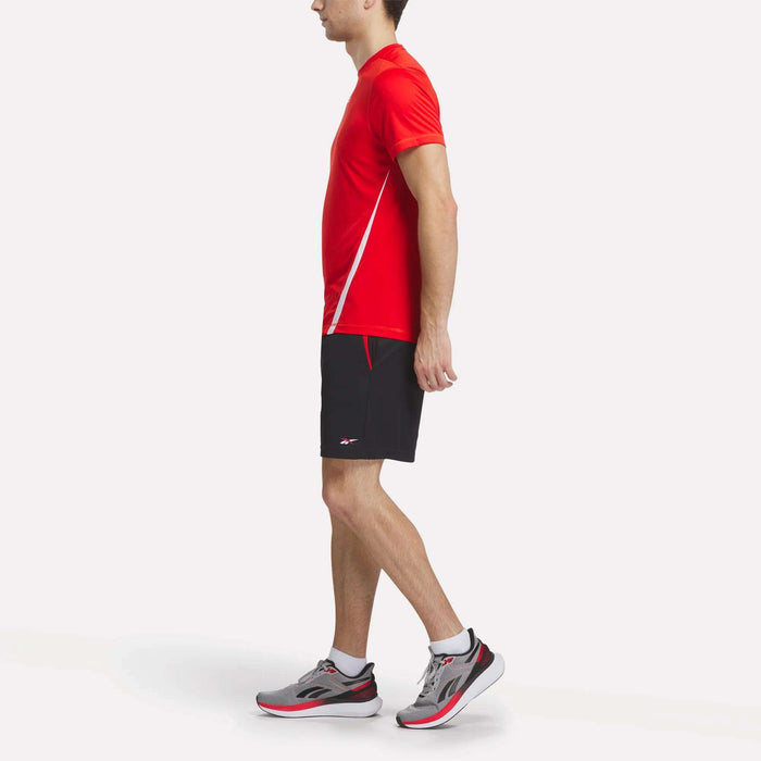 A man wearing a red athletic shirt, Reebok Men's Train Blocked Knit Shorts in black, and gray running shoes stands in profile against a plain white background.