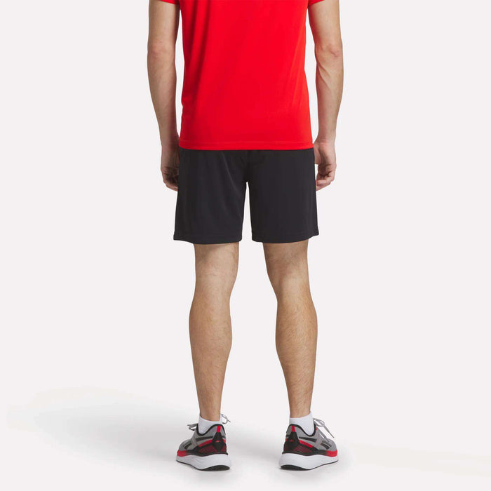 A person faces away, wearing a bright red shirt and Reebok Men's Train Blocked Knit Shorts, along with gray athletic shoes featuring red and black accents, all set against a plain white background.
