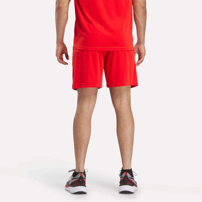 A person stands facing away from the camera, wearing a red T-shirt, Reebok Men's Train Blocked Knit Shorts, and black, white, and red sneakers against a plain white background—a stylish workout look.