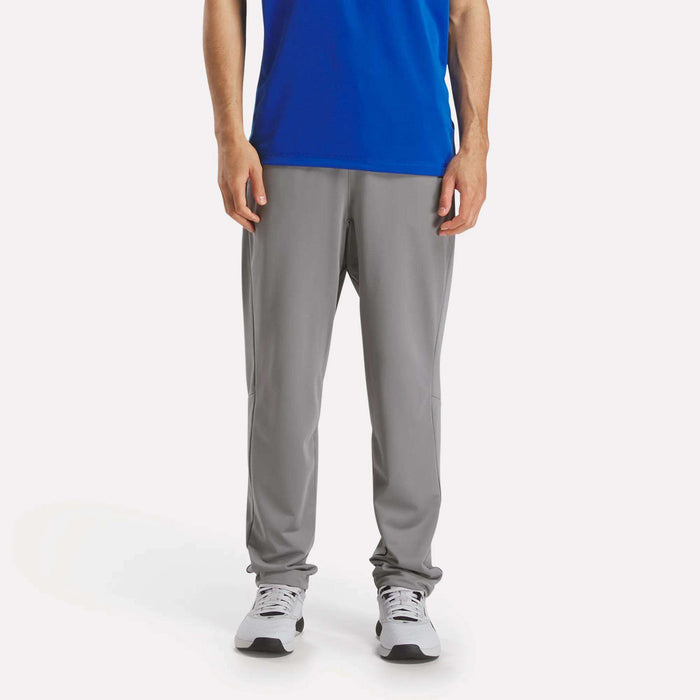 A person in a blue shirt and Reebok Men's Train Knit Pants in light gray, paired with white sneakers, stands facing forward against a plain white background. The image is cropped at the shoulders.