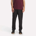 Against a plain white background, a person wears Reebok Men's Train Knit Pants with black sneakers and a maroon short-sleeve shirt. The photo, cropped at the shoulders, focuses on the lower half of the body.