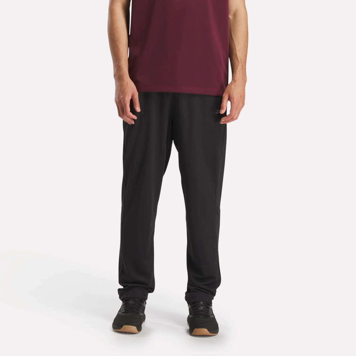 Against a plain white background, a person wears Reebok Men's Train Knit Pants with black sneakers and a maroon short-sleeve shirt. The photo, cropped at the shoulders, focuses on the lower half of the body.