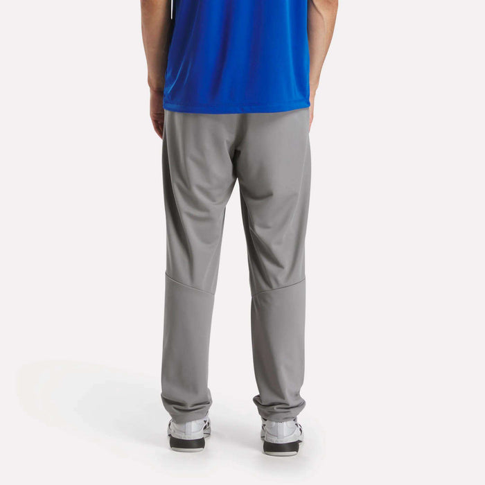 A person in a blue shirt, white sneakers, and Reebok Men's Train Knit Pants in gray stands with their back to the camera against a plain white background.