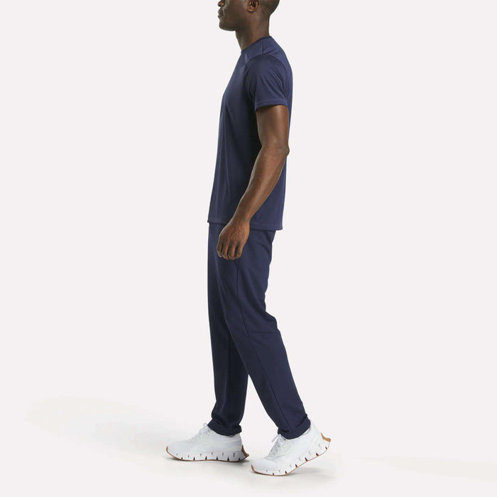 A man stands sideways wearing Reebok Men's Train Knit Pants in navy blue with a matching T-shirt and white athletic shoes, set against a plain white background.