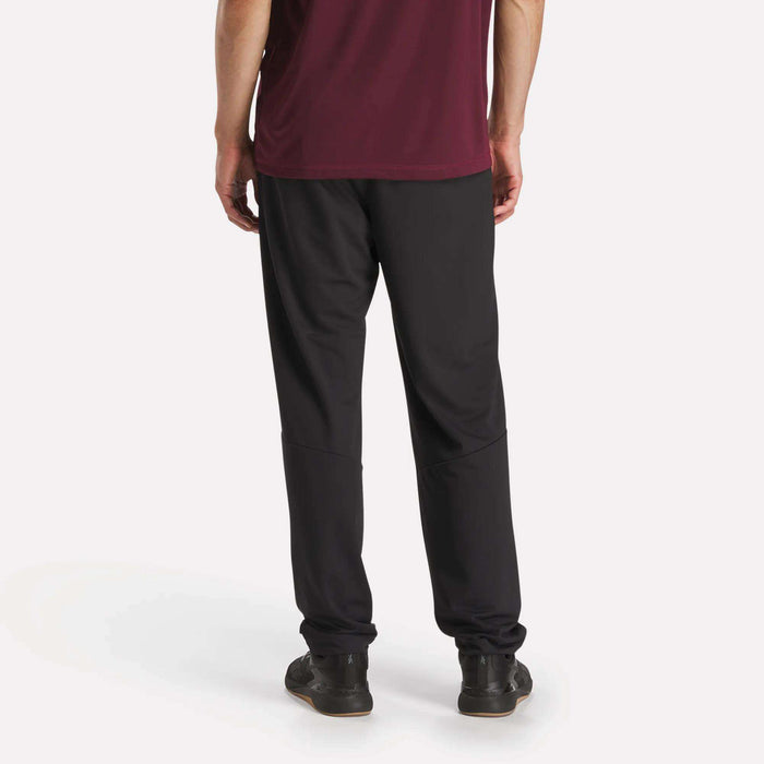 A person stands with their back to the camera, wearing a maroon shirt and black Reebok Men's Train Knit Pants with black sneakers, set against a plain white background.