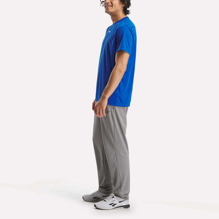 A man stands sideways, smiling, wearing a bright blue short-sleeve shirt, Reebok Men's Train Knit Pants in light gray, and white athletic shoes against a plain white background.