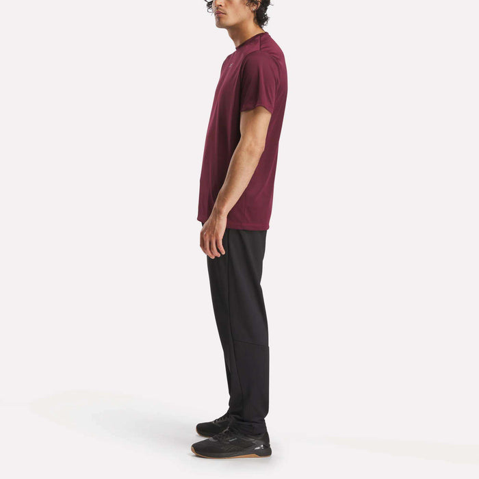 A man stands in profile wearing a burgundy t-shirt, black Reebok Men's Train Knit Pants, and black sneakers against a plain white background.