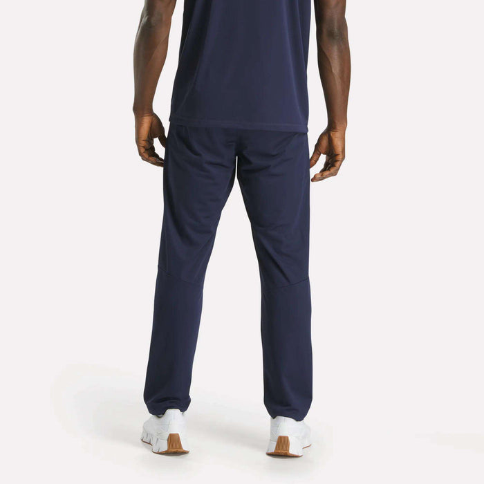 A man faces away, wearing a navy blue T-shirt and Reebok Men's Train Knit Pants with white sneakers on a plain light background.