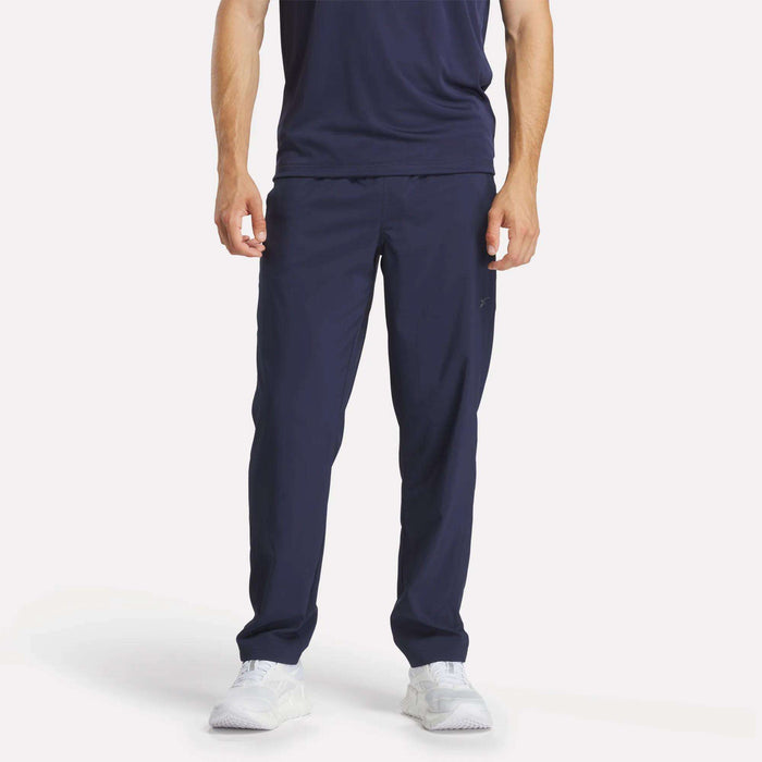 A person stands facing forward against a plain light background, wearing Reebok Men's Train Woven Unlined Pants in navy blue with a matching t-shirt and white athletic shoes. The person's head is not visible in the image.